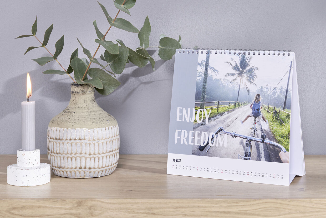 Une table avec un calendrier illustrant une personne à vélo sur une route bordée d'arbres. Une bougie et des décorations à côté.