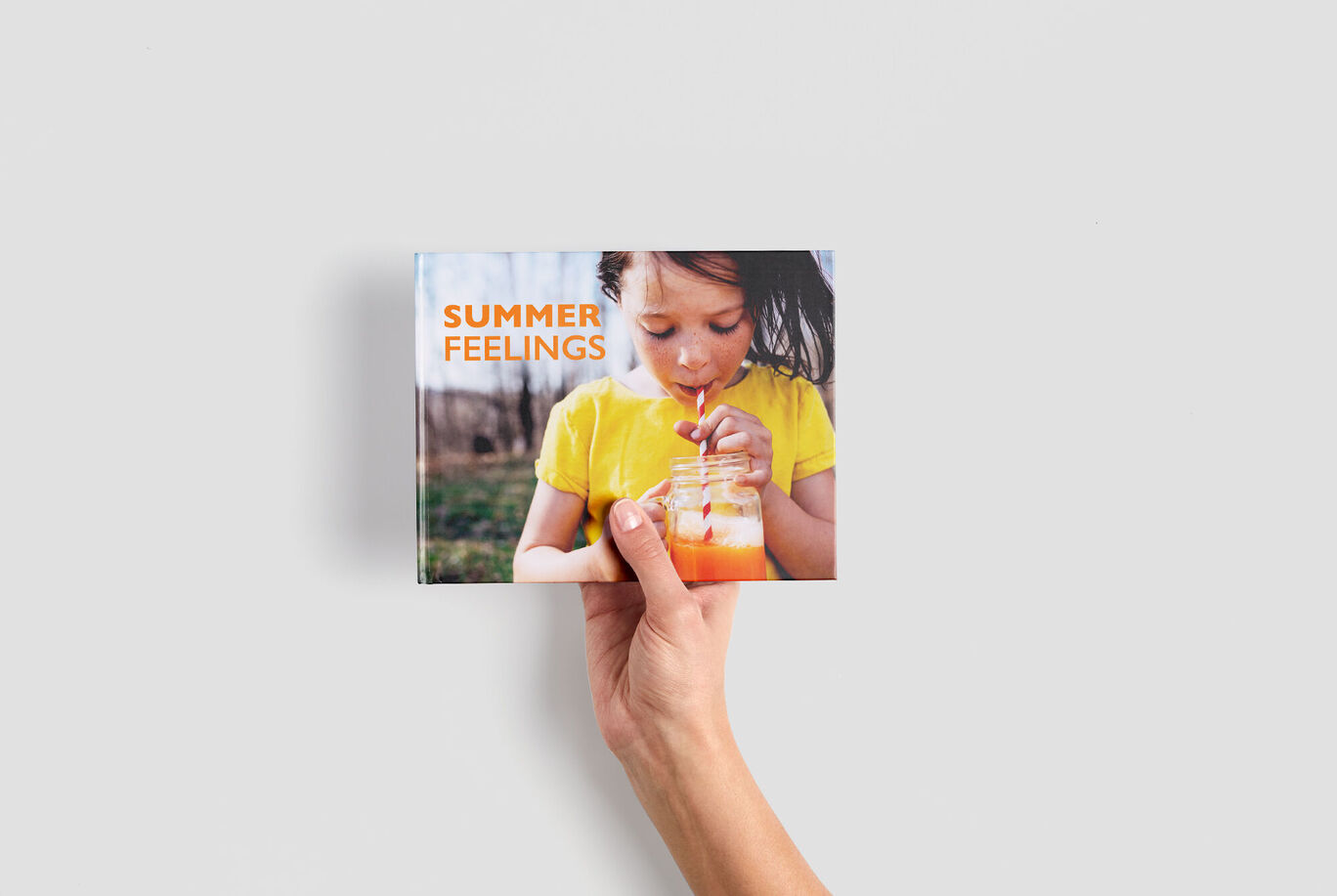 Une main tient un petit livre photo au format paysage avec une couverture estivale et le texte Summer Feelings