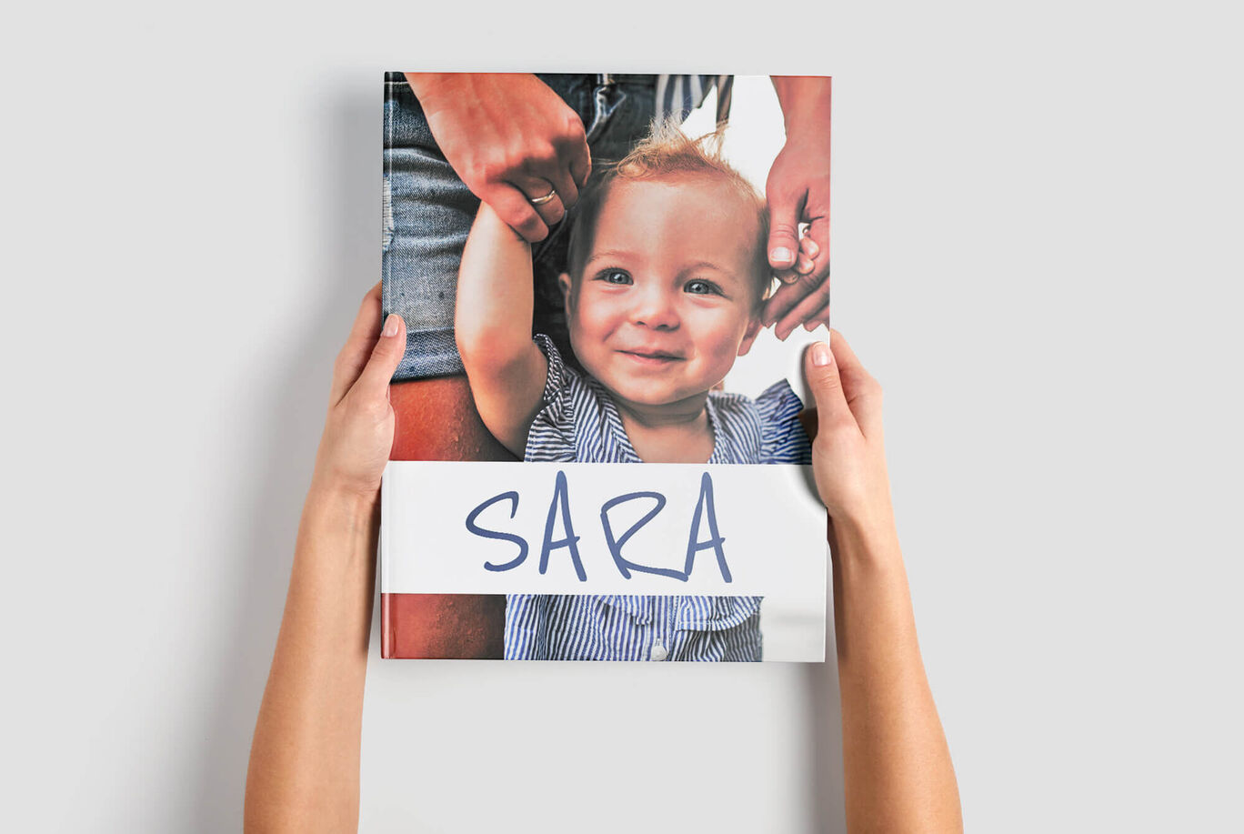Deux mains tiennent un livre photo avec le prénom Sara en couverture et la photo d’un enfant tenu par la main