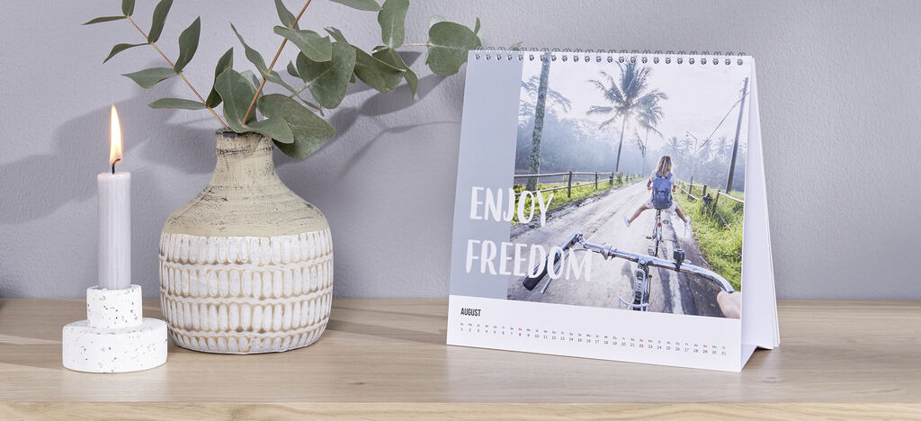Un magnifique calendrier de table avec une photo d'une fille sur une route bordée d'arbres et une branche d'eucalyptus en arrière-plan. Le calendrier affiche le texte 'ENJOY FREEDOM' dans une ambiance chaleureuse et accueillante.