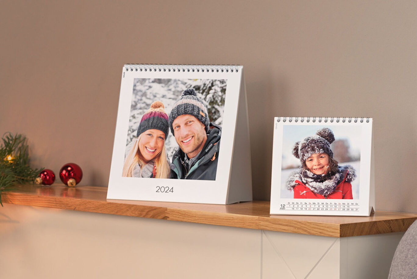 Twee mooie CEWE fotokalenders op een houten plank. De grote toont een winterlandschap, de kleine heeft een rood jasfoto.