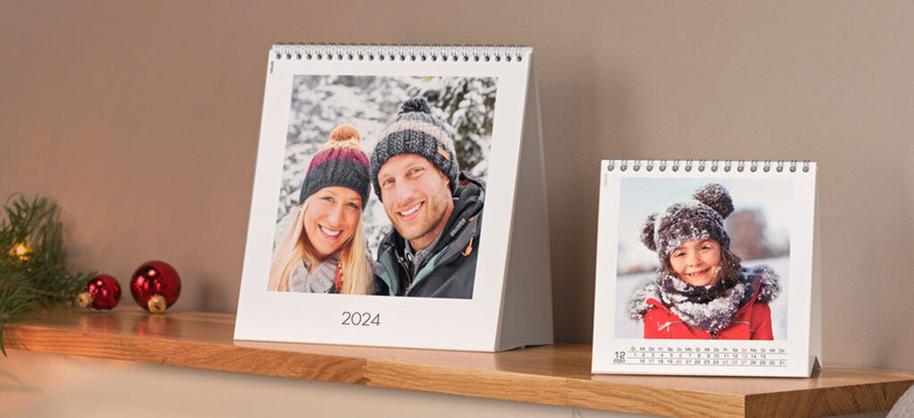 Twee gepersonaliseerde fotokalenders, een groot en een klein, op een houten plank. Beide tonen winterbeelden en vreugdevolle momenten.
